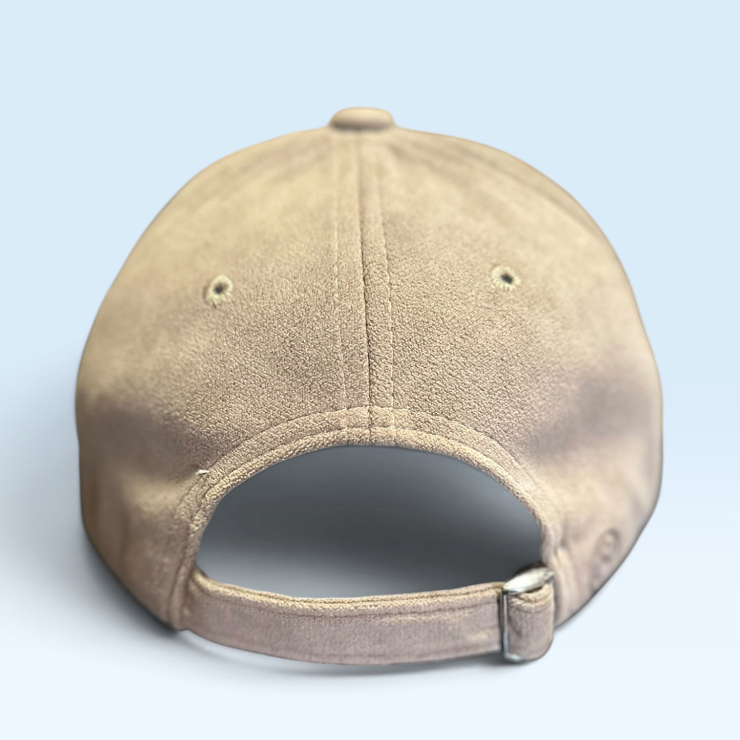 A beige baseball cap shown from the back, featuring a textured suede-like finish, ventilation eyelets, and an adjustable strap closure, set against a light blue background.
