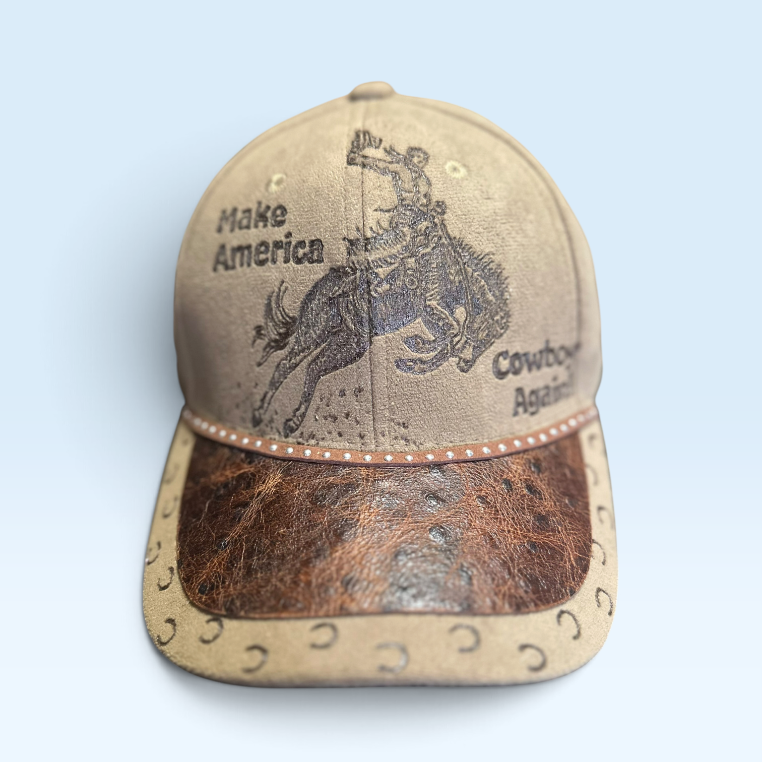 A western-style baseball cap with a tan leather crown, a brown ostrich-embossed visor, and black printed graphics of a cowboy on a bucking horse with the text 'Make America Cowboy Again'.