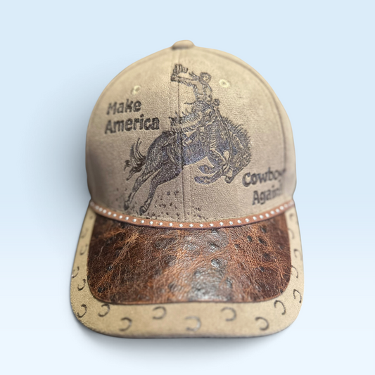 A western-style baseball cap with a tan leather crown, a brown ostrich-embossed visor, and black printed graphics of a cowboy on a bucking horse with the text 'Make America Cowboy Again'.