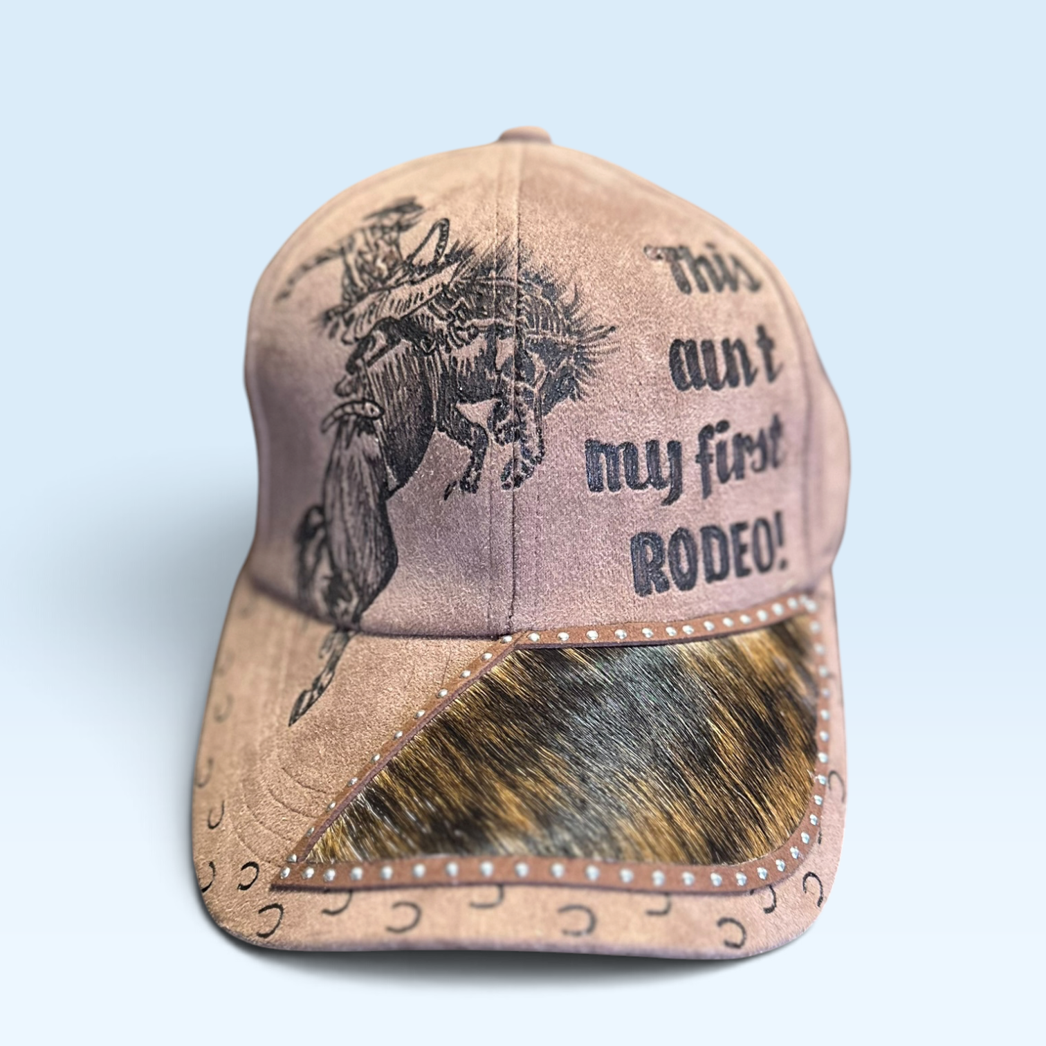 A brown baseball cap with a western theme, featuring a hand-burned graphic of a cowboy riding a bucking bronco on the front, the text 'This ain't my first RODEO!' on the side, and real brown and black brindle cowhide on the brim.