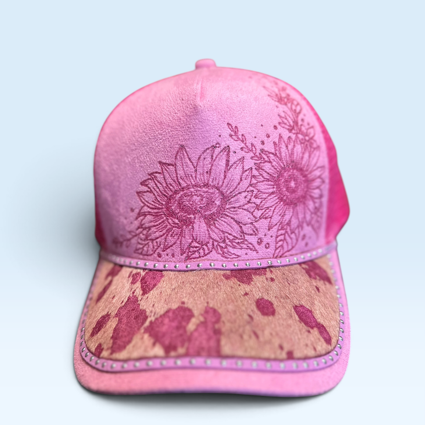 A pink trucker-style baseball cap with a suede-like front panel, floral sunflower print, and a distressed tan brim with pink splatter details, shown against a light blue background.