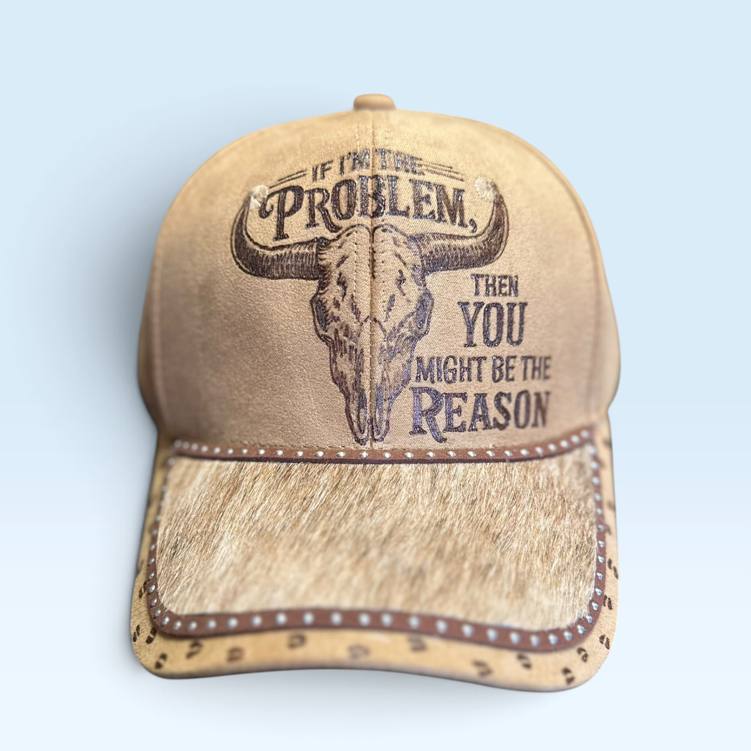 A tan vegan suede baseball cap with a western design, featuring a skull graphic and the text 'IF I'M THE PROBLEM, THEN YOU MIGHT BE THE REASON' on the front, featuring a brindle hair on hide leather brim set against a light blue background.
