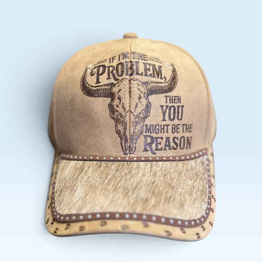 A tan vegan suede baseball cap with a western design, featuring a skull graphic and the text 'IF I'M THE PROBLEM, THEN YOU MIGHT BE THE REASON' on the front, featuring a brindle hair on hide leather brim set against a light blue background.