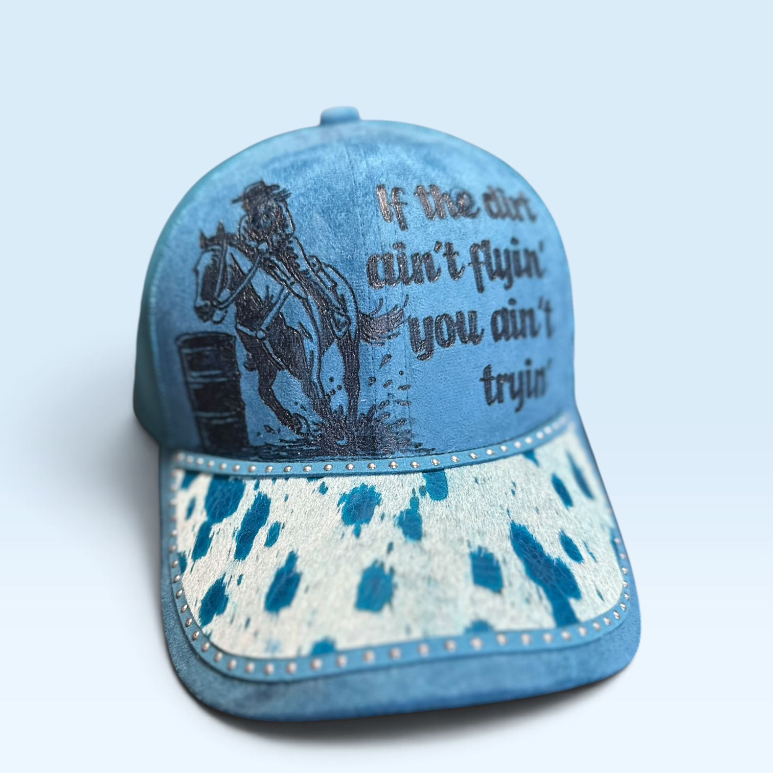A teal blue baseball cap with a cowhide-patterned visor, featuring a black graphic of a cowgirl barrel racer on a horse and the text 'If the dirt ain't flyin' you ain't tryin''.