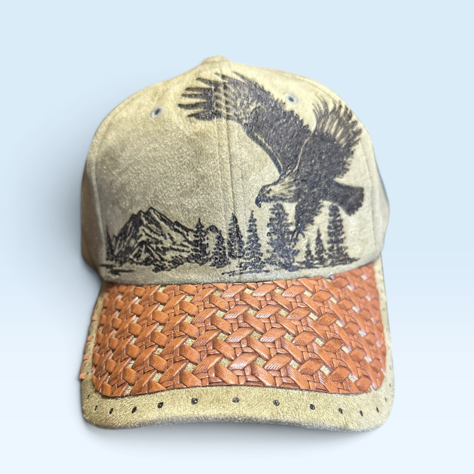 A baseball cap featuring a moss green vegan suede crown with a black burned eagle and mountain landscape design, a brown woven leather brim, and a light blue background.