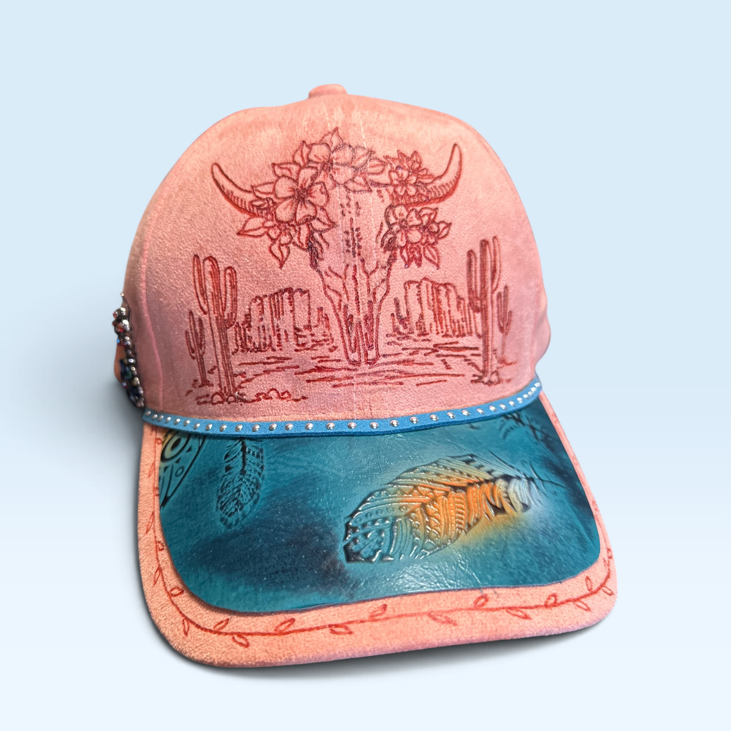 A western-style baseball cap with a dusty pink (light terra cotta) crown, featuring an hand burned desert landscape with a skull and cacti on the front. The curved brim is features turquoise leather with a feather design and a small coral accent.