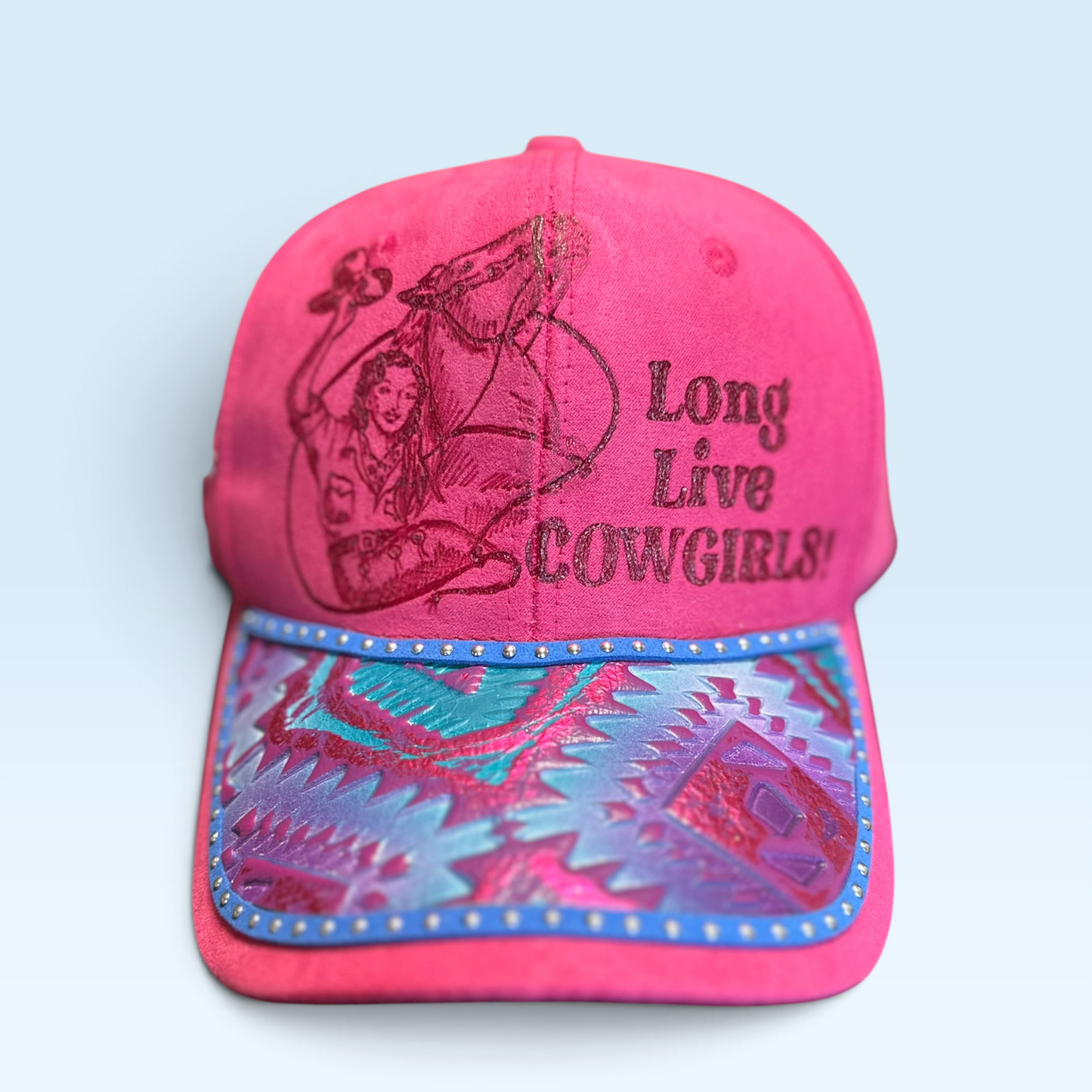 A bright pink baseball cap featuring hand burned artwork and text on the front panel, with a patterned brim in pink, teal, and blue, set against a light blue background.