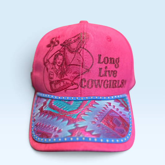 A bright pink baseball cap featuring hand burned artwork and text on the front panel, with a patterned brim in pink, teal, and blue, set against a light blue background.