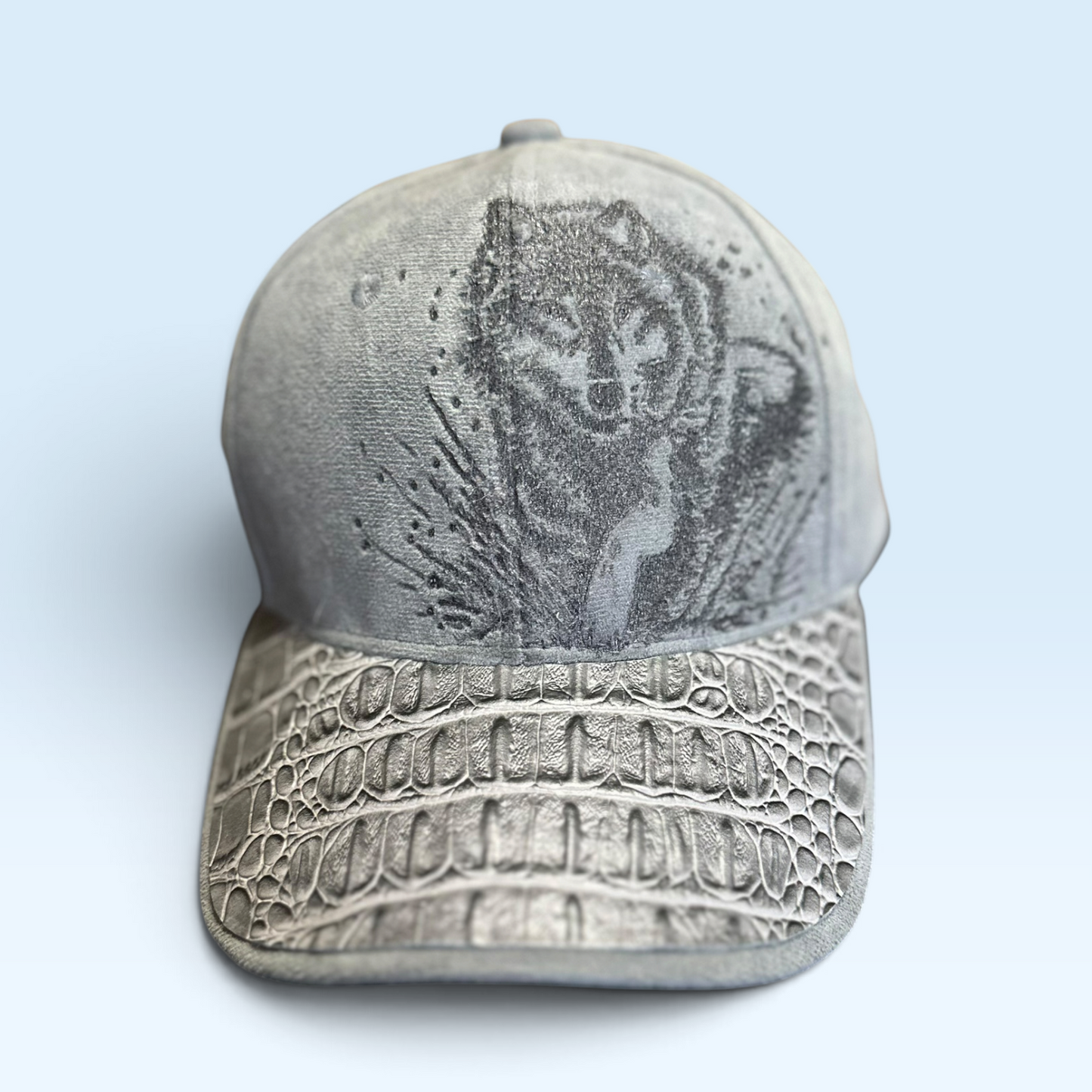 A distressed gray baseball cap featuring a crocodile-embossed brim and a graphic of a wolf on the front, set against a light blue background.