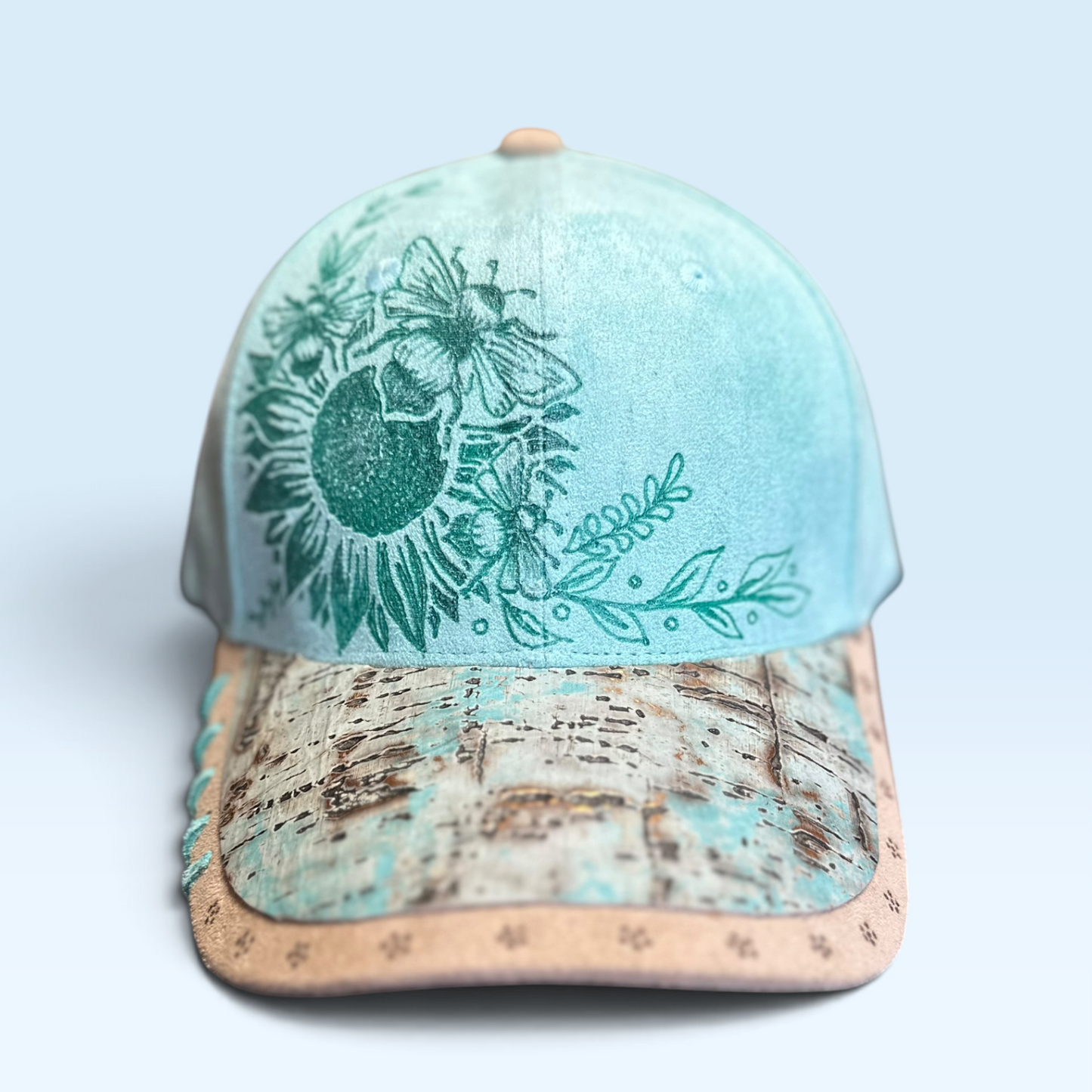 A baseball cap with a turquoise and beige crown, featuring a green floral sunflower graphic with bees on the front panel and a leather cork-textured visor.