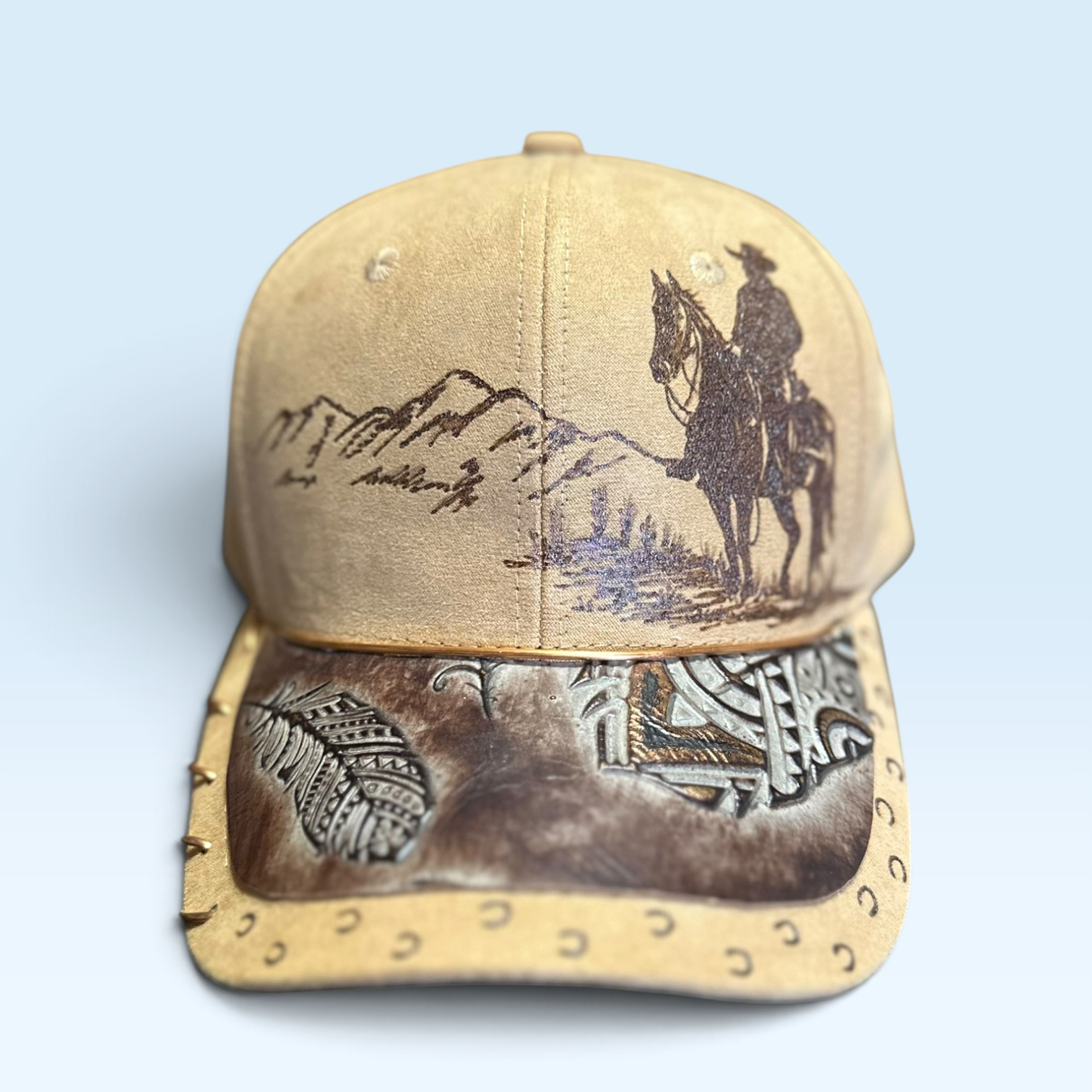 A light brown western-style baseball cap featuring a brown leather brim with decorative stitching and a western-themed graphic print. The front of the cap is light brown suede-like material with a dark brown mountain and cowboy on horseback design.