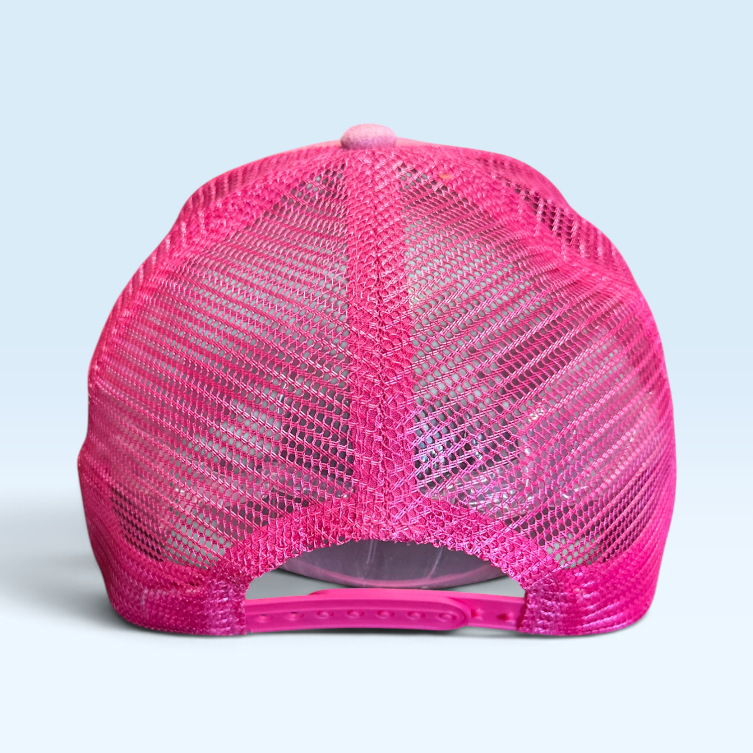A bright pink trucker-style cap shown from the back, featuring a pink mesh back and a matching pink plastic snap closure, set against a solid light blue background.