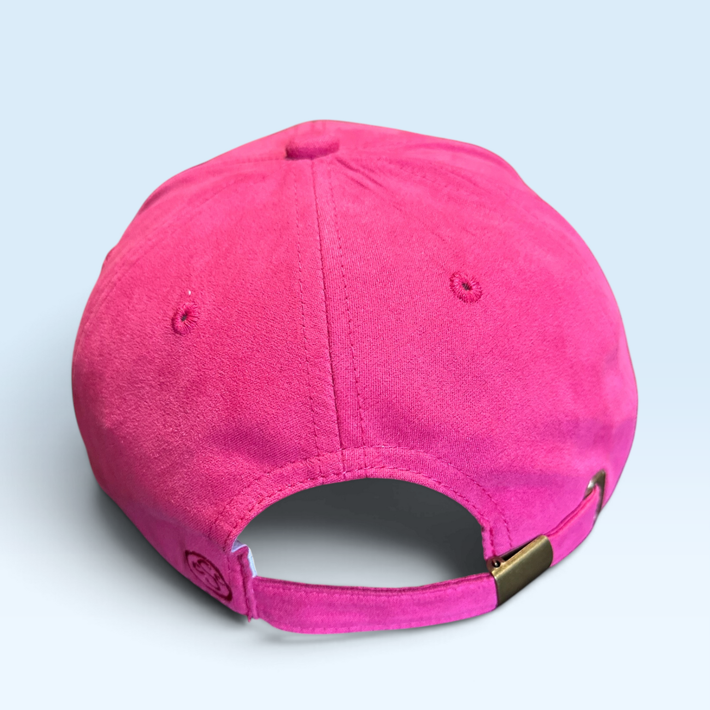 A bright pink baseball cap shown from the back, featuring a fabric texture and an adjustable strap with a small gold metal accent, set against a light blue background.