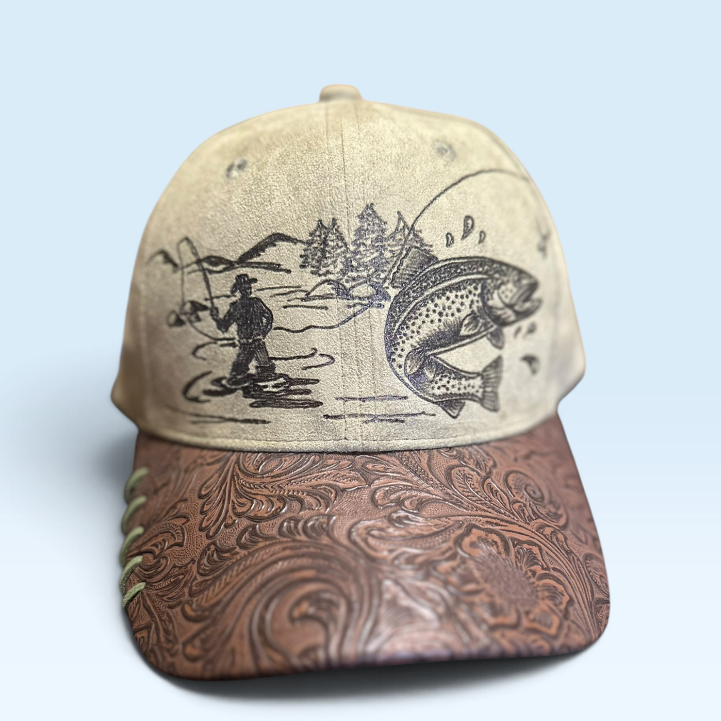 A baseball cap with a beige fabric crown featuring a black fishing illustration, and a brown tooled leather brim with a swirling pattern, set against a light blue background.